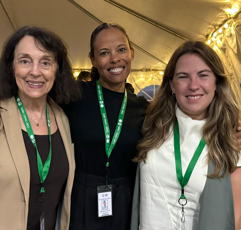 Three women stand together smiling under a tent, each wearing a green lanyard with an ID badge. There are string lights in the background
