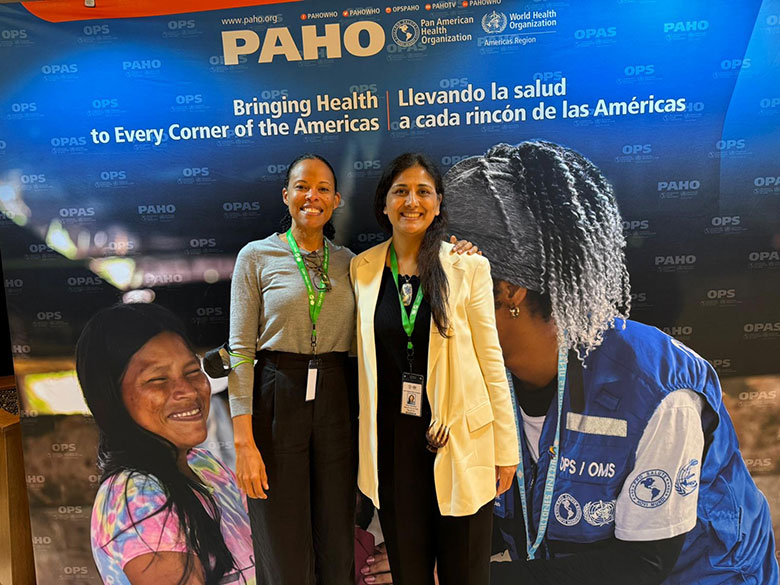 Two women smile and stand side by side in front of a colorful PAHO/WHO banner promoting health across the Americas, featuring images of healthcare workers and a smiling woman in traditional attire