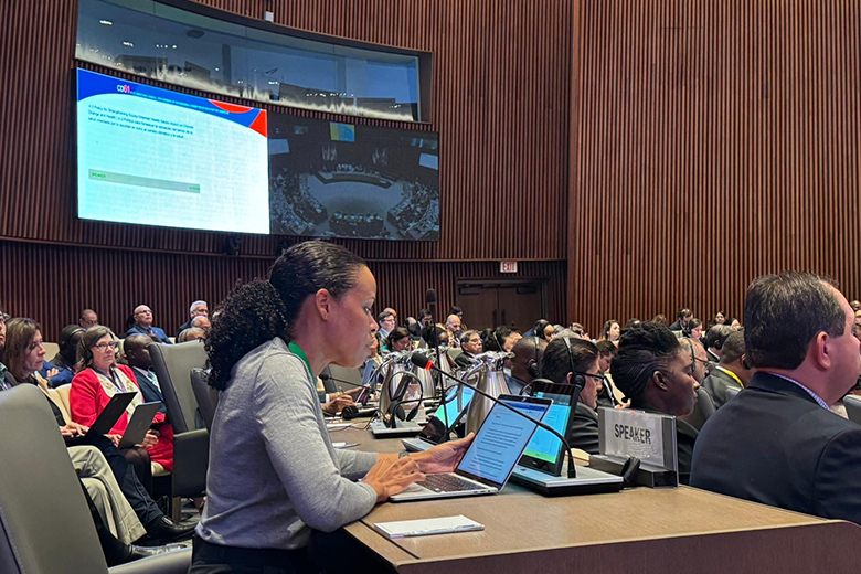 People sit in a large conference room, many with laptops open. A woman at the front speaks into a microphone. Behind her, a screen displays a presentation and there are monitors showing a view of the room