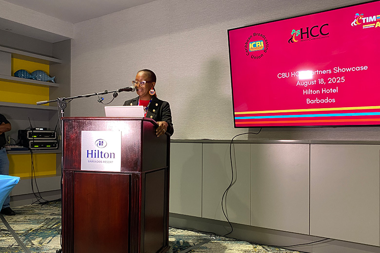 Sheena Warner-Edwards presenting at a podium with a large tv screen behind her
