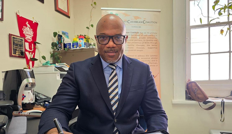 A bald headed Caribbean man wearing spectacles sitting at an office desk signing a document