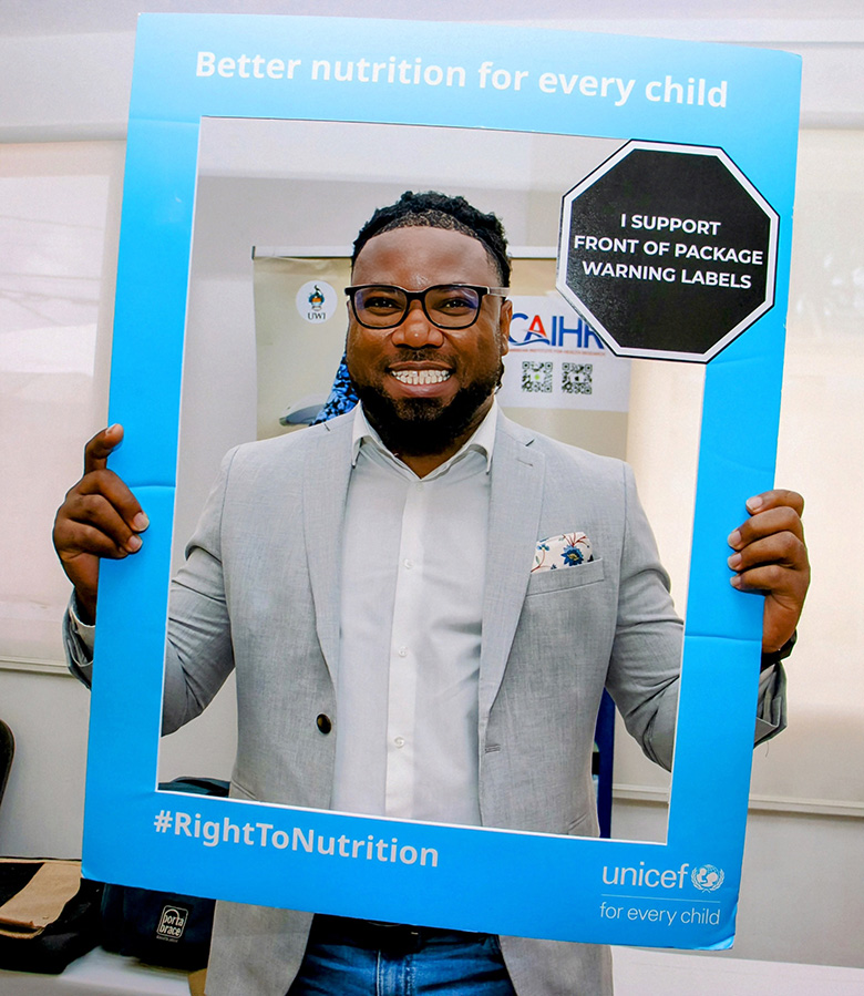 Smartly dressed man holding a blue picture frame with Better Nutrition for every child written on the top of the frame with an octagonal black shape at the top right of the fremae with the words I support front of package warning labels in bold lettering