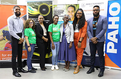 Representatives from PAHO, UNICEF, and Ross University pose together at a collaborative health event. The group is positioned against a row of informative banners that highlight regional health initiatives. The individuals are arranged in a line, showcasing a mix of professional office wear and branded event t-shirts in a well-lit indoor setting.