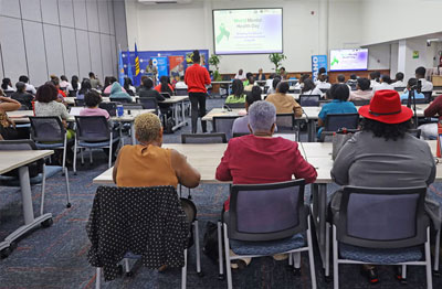 An indoor seminar or workshop setting dedicated to World Mental Health Day. The room is filled with participants seated in rows, looking toward a lit projector screen and a panel of speakers at the front. To the left of the stage area, various international and organisational flags are displayed alongside promotional materials. The atmosphere is professional and focused, with the audience engaged in the presentation.