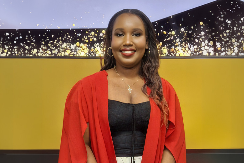 A smiling woman in a red cape jacket and black top, standing before a yellow background with a glittering gold display at the top