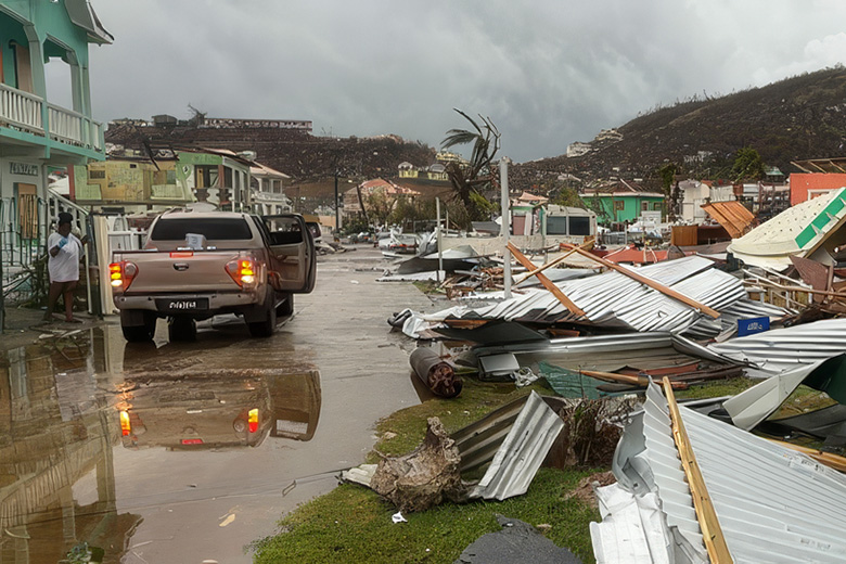 Destruction on Union Island after Hurricane Beryl, showing a flooded road, a brown pickup truck, and a person standing amidst heavy debris. The street is littered with collapsed metal roofing and shattered building materials beneath a gloomy, overcast sky.