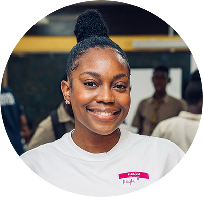 Circular headshot of a smiling young Black woman wearing a white shirt with a pink 'Hello my name is Kayla' name tag. The background features a softly out-of-focus indoor group setting.
