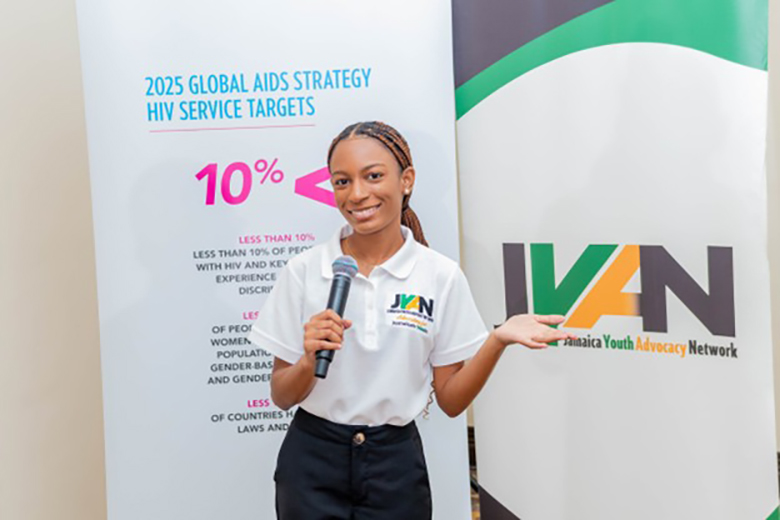 A woman wearing a white JYAN polo shirt holding a microphone and gesturing toward a Jamaica Youth Advocacy Network banner. To her left is an educational banner detailing the 2025 Global AIDS Strategy and HIV service targets.