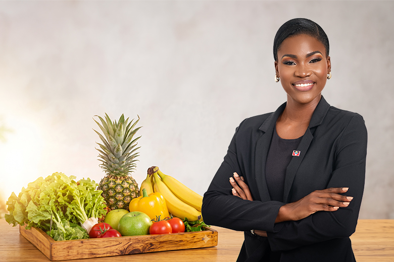 Smiling woman in a black jacket standing next to a wooden display of vibrant, healthy fruits and vegetables.