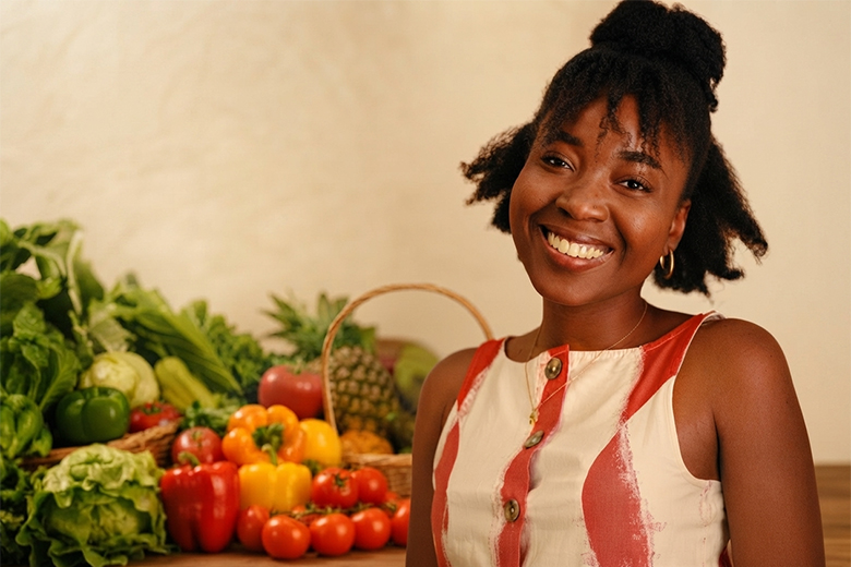 Close-up of a smiling woman in a red and white top, with a colourful assortment of fresh produce and a fruit basket in the background