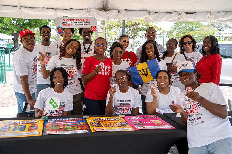 A group of campaigners gathered under a marquee advocating against unhealthy food marketing in schools, holding signs and a Barbados flag