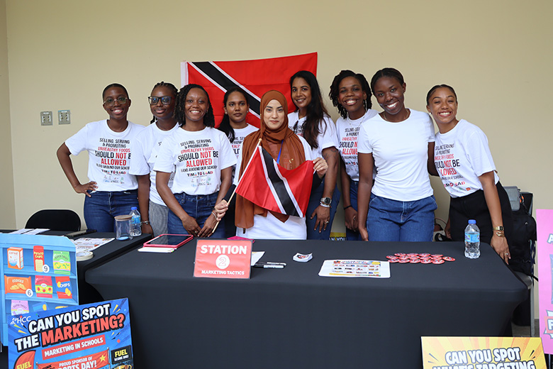 Campaigners holding Trinidad and Tobago flags pose behind an information table with posters about healthy food marketing