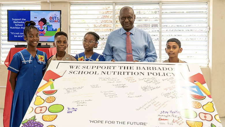 Four students and a smiling man in a blue shirt stand behind a signed poster supporting the Barbados School Nutrition Policy