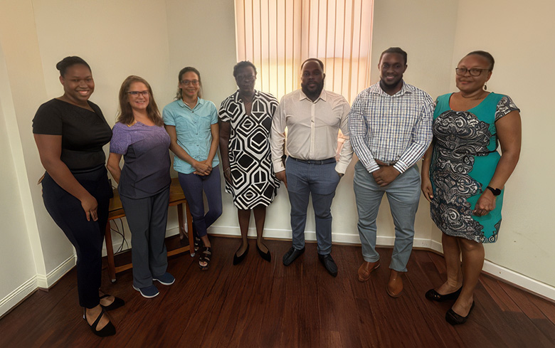 Seven colleagues stand side by side in a bright office room, posing together for a group photograph