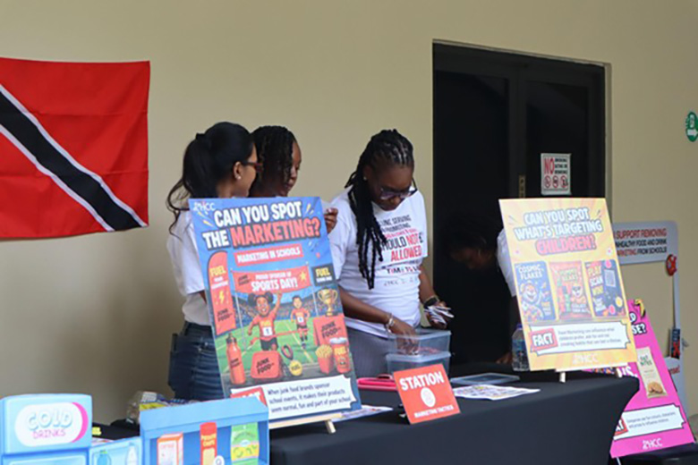 Three people manage an event table with posters about food marketing to children and a Trinidad and Tobago flag on the wall