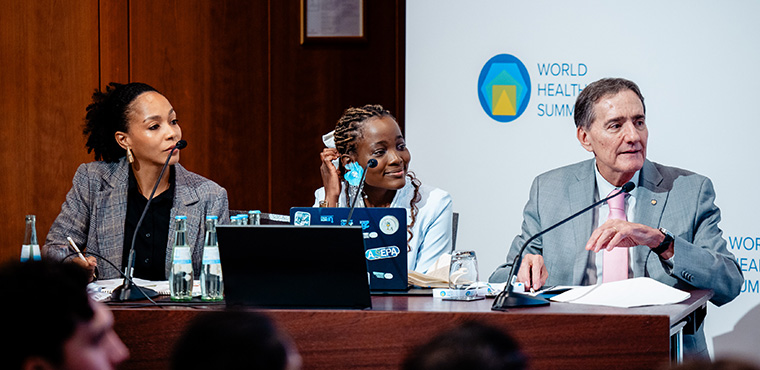 A panel discussion at the World Health Summit featuring two women and a man speaking with laptops and microphones on their desk