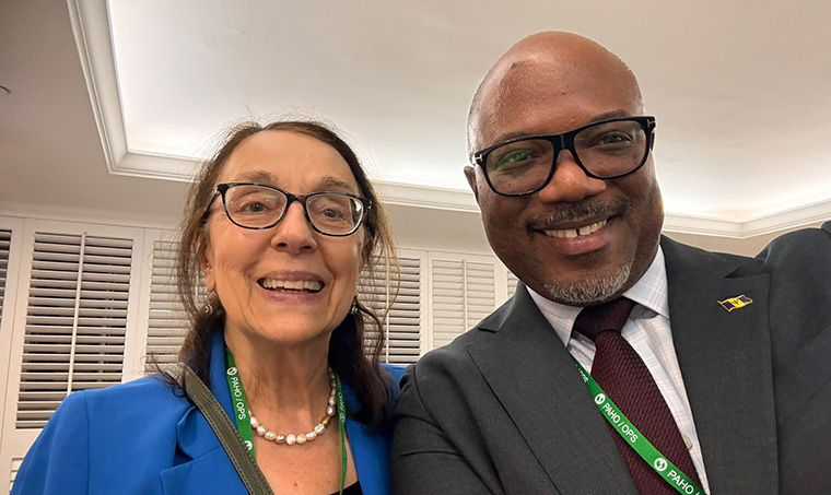 Two smiling delegates wearing PAHO/OPS lanyards; a woman with glasses and a pearl necklace alongside a man in a dark suit and Barbados flag pin