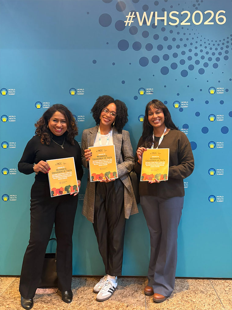 hree women holding yellow HCC report booklets stand in front of a blue World Health Summit backdrop display featuring the hashtag WHS2026