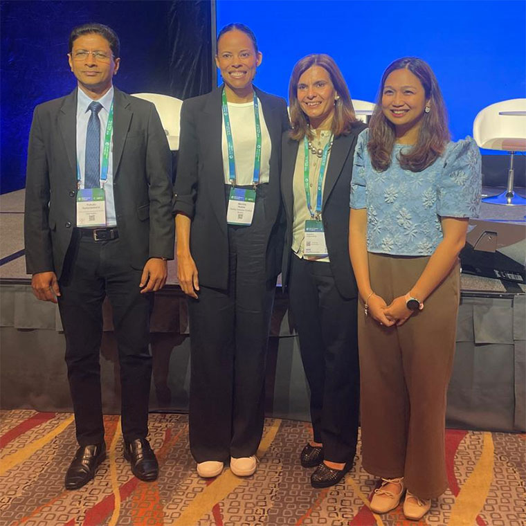 Four smartly dressed speakers standing side by side on a stage, three wearing event badges around their necks