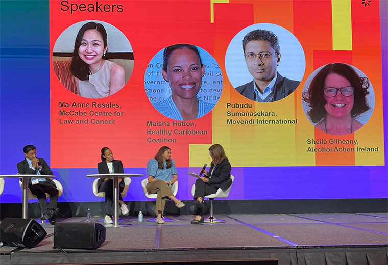 Panel discussion on stage with four speakers seated beneath a backdrop showing headshots of the featured panellists